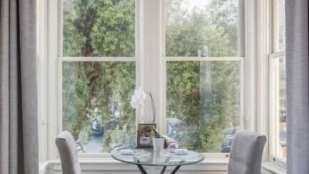 Ein Zimmer mit großem Fenster, zwei Stühlen, einem kleinen runden Tisch und einem Bett.