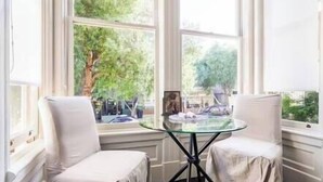Ein Zimmer mit zwei weißen Stühlen, einem Glastisch und großen Fenstern mit Blick auf Bäume und eine Straße.
