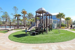 A playground with a slide and climbing structures, surrounded by a well-maintained lawn and palm trees.