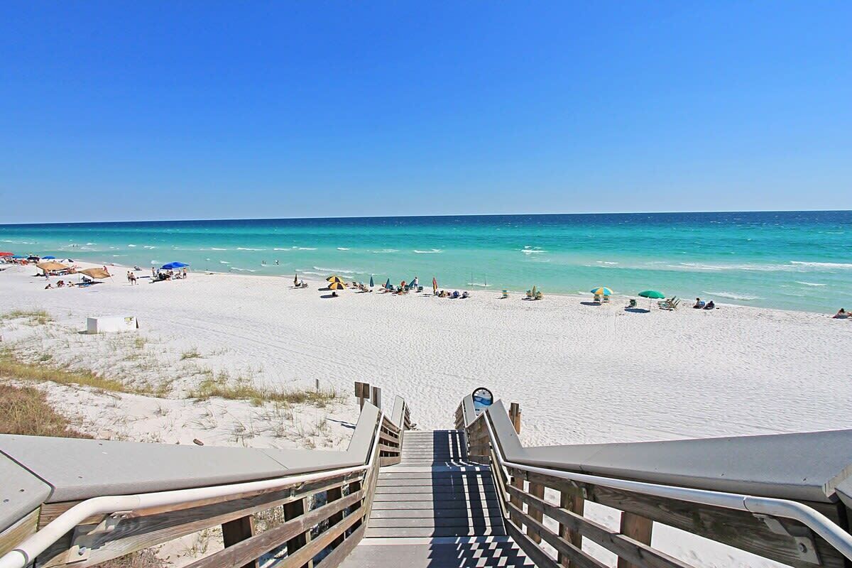 A beach with a wooden staircase leading down to the sand, a clear blue sky, and the ocean in the background.