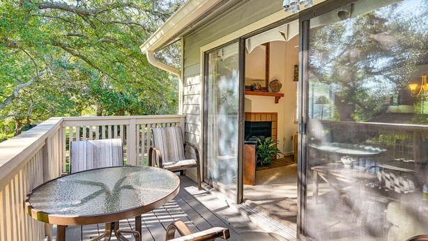 Een overdekt terras buiten, met een glazen tafel en stoelen, een houten terras en uitzicht op het interieur door een glazen deur.