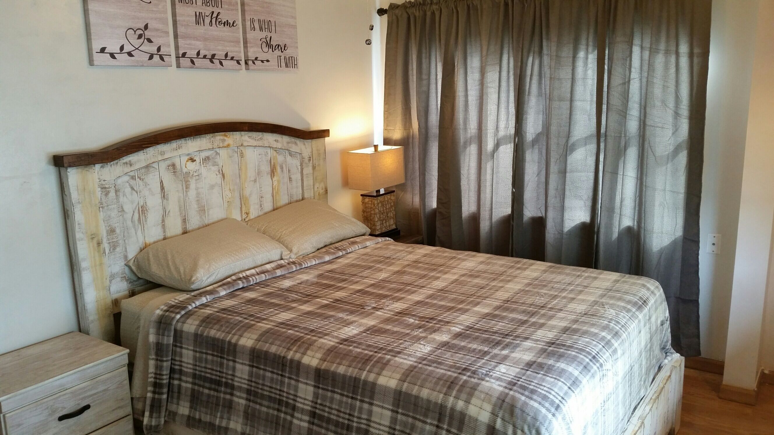 Een slaapkamer met een houten hoofdbord, een geruite sprei, een bedlampje en een raam met gordijnen.