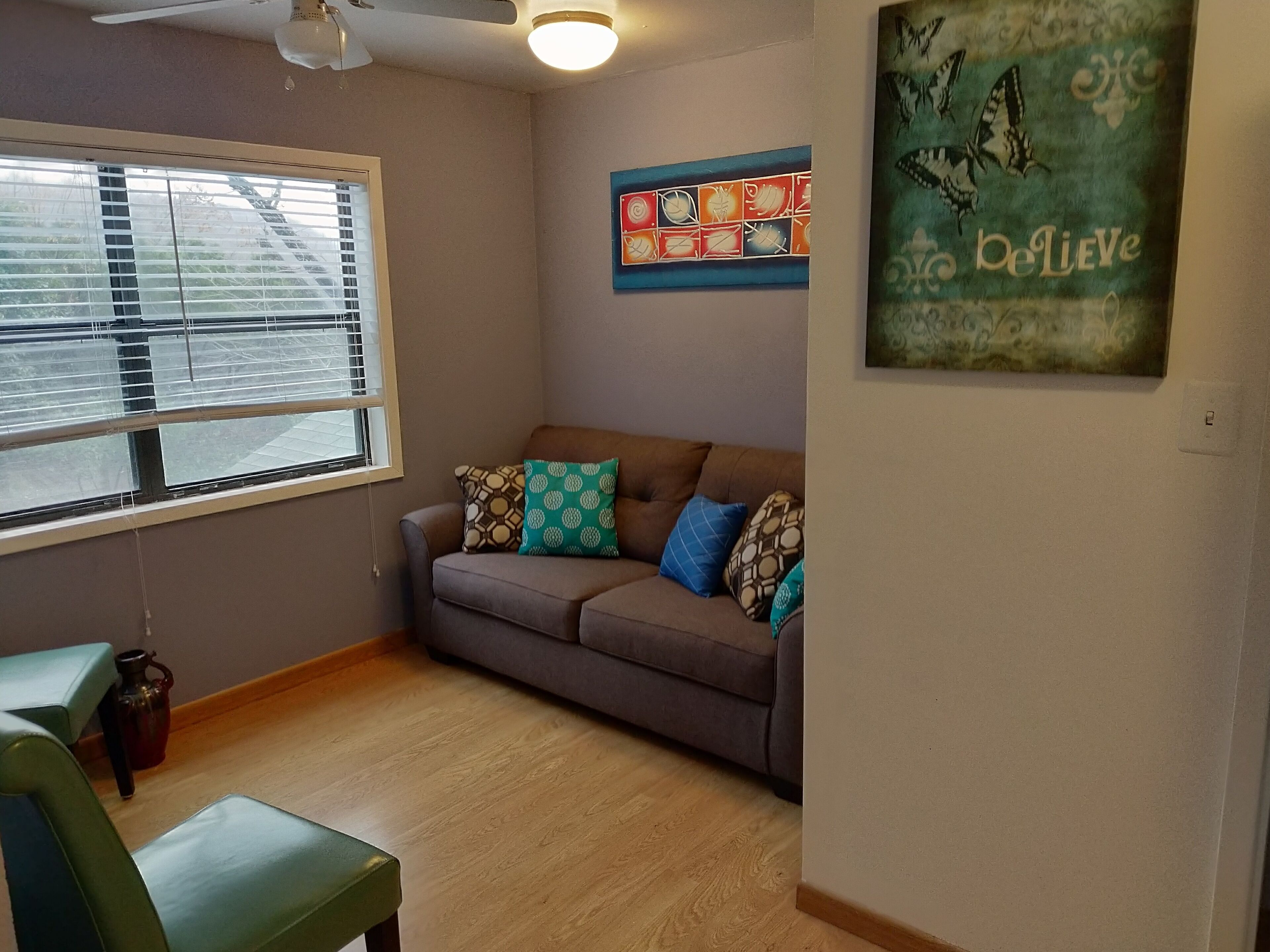Ein Wohnzimmer mit einem braunen Sofa, gemusterten Kissen, einem Fenster mit Jalousien, einem Deckenventilator und einer Wand mit einem Schmetterlingsbild und einem motivierenden Zitat.