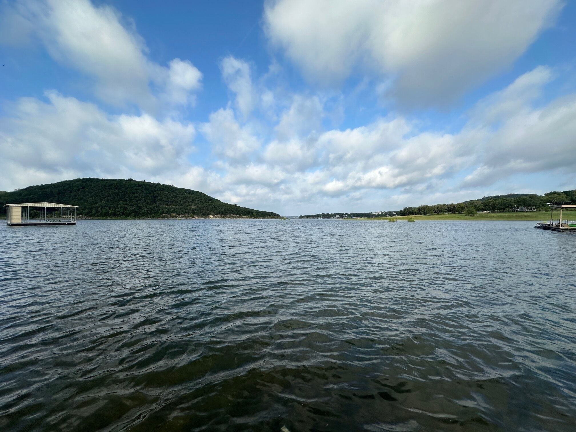 En sø med en anløbsbro og et overdækket område, omgivet af bakker og en delvist overskyet himmel.