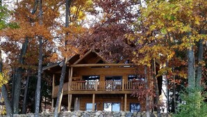 Een chalet met een stenen muur en een houten terras, omgeven door bomen met herfstbladeren.