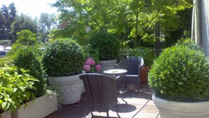 Eine Terrasse im Freien mit einem runden Tisch und zwei Korbstühlen, umgeben von großen Kübelpflanzen und verschiedenen Bäumen.