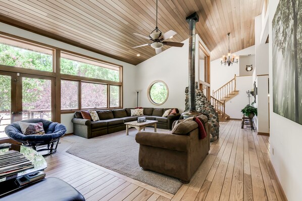 A spacious living room with a stone fireplace, large windows, a ceiling fan, and a staircase leading to an upper level. - A river runs through. Private Estate on OAK CREEK! Pickleball Court in backyard! (Sedona)