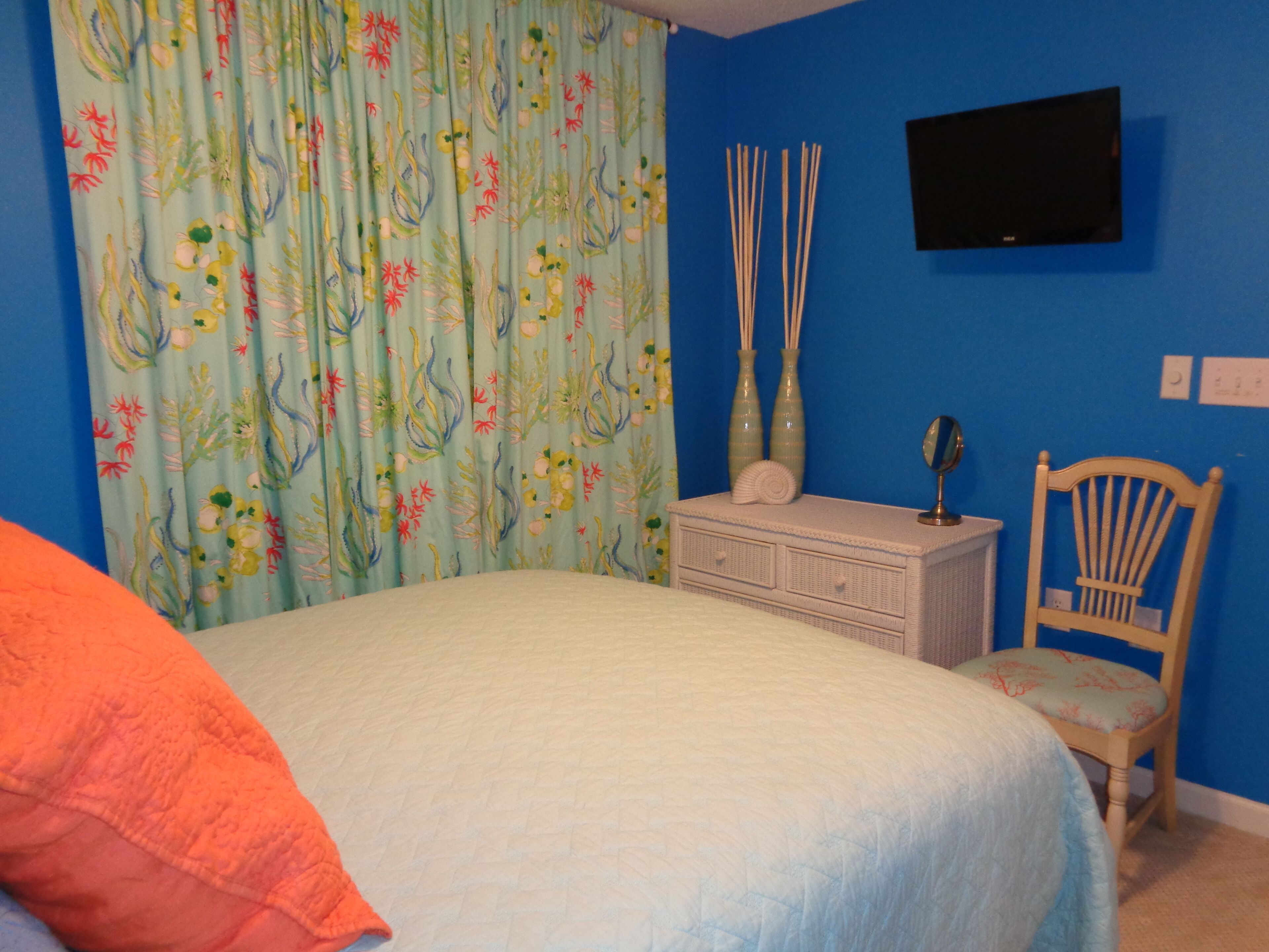 A bedroom with a bed, a dresser, a chair, a television on the wall, and a curtain with a floral pattern.