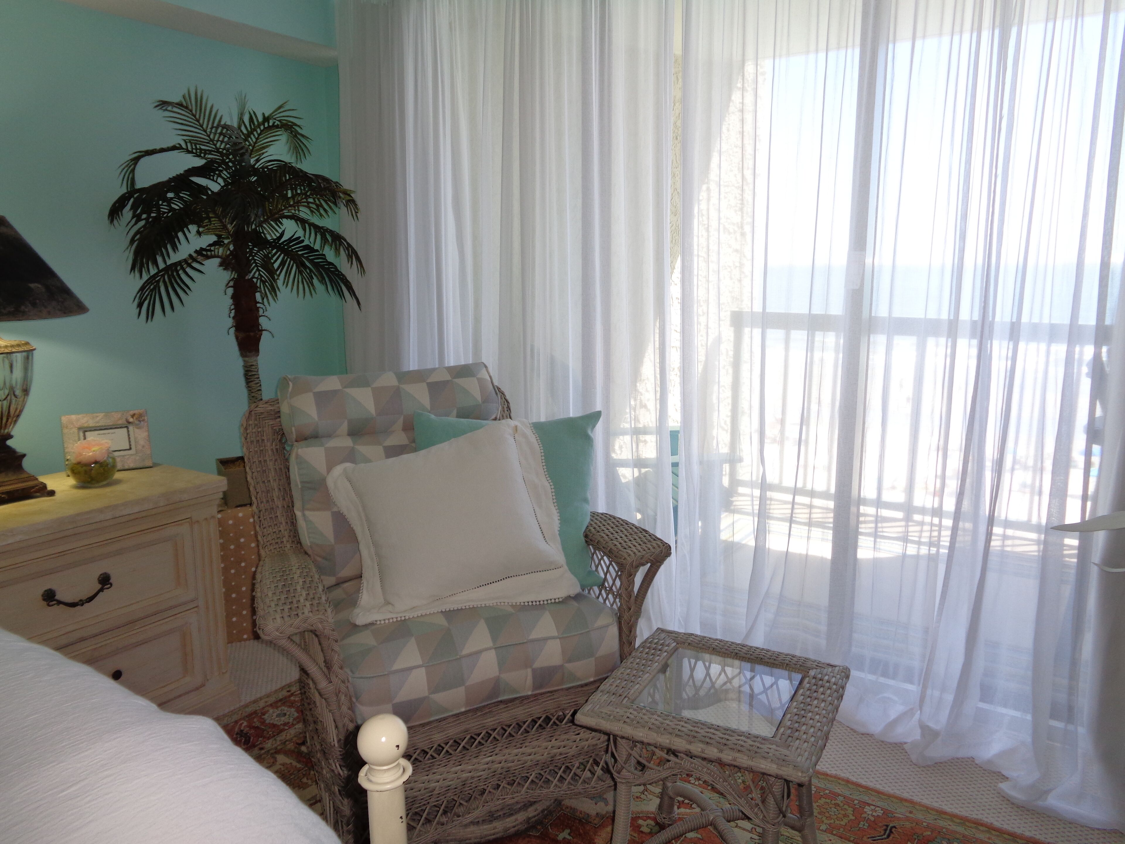 A room with a bed, a chair with a geometric cushion, a small table, a lamp, a plant, and a window with sheer curtains.