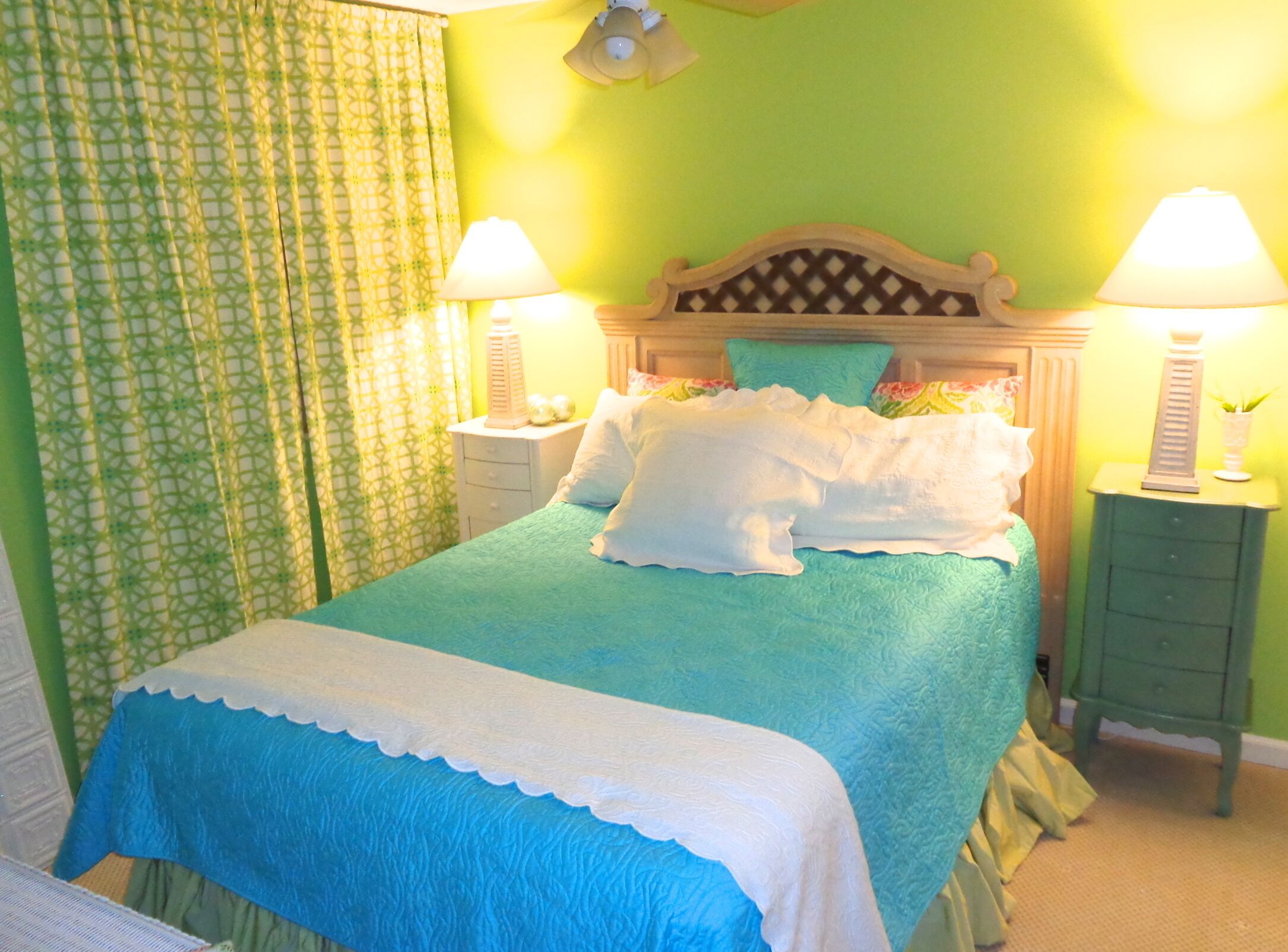 A bed with a blue bedspread and white pillows, two bedside tables with lamps, and a green patterned curtain.