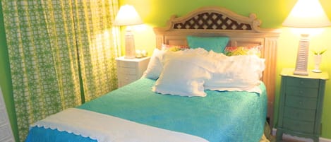 A bed with a blue bedspread and white pillows, two bedside tables with lamps, and a green patterned curtain.
