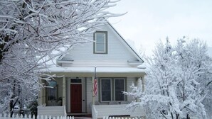Una casa cubierta de nieve con un porche delantero, una puerta roja y una bandera estadounidense.