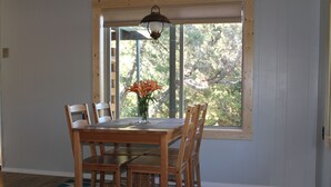 A dining area with a wooden table and chairs, a vase with flowers, and a window with a view of trees.