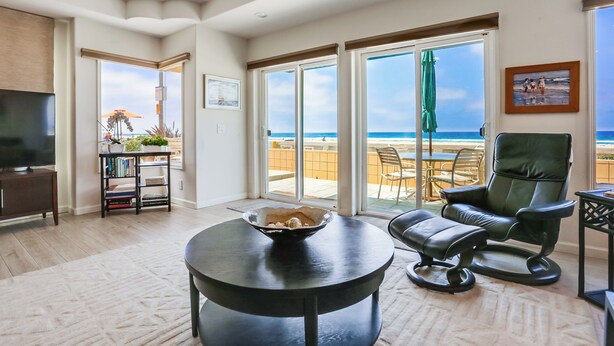 A spacious living room with a large window offering a view of the beach, a comfortable leather chair, a small round table with a bowl, and a bookshelf. - Amazing ocean front, ground floor condo in Mission Beach! Book now and save! (San Diego)