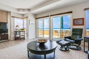 Een zithoek met een groot raam met uitzicht op een strand, een zwarte leren fauteuil, een ronde tafel met een schaal en een boekenkast.