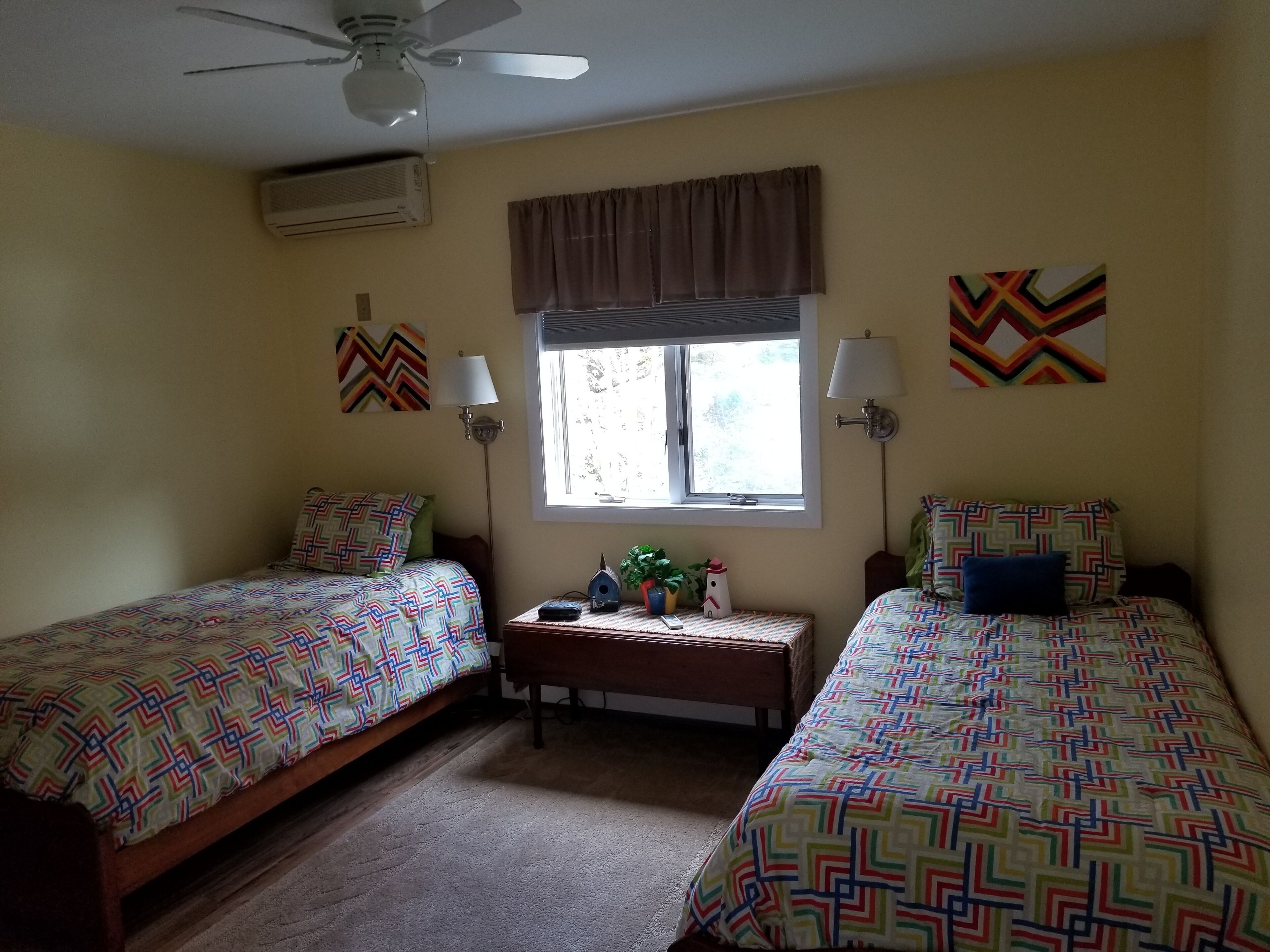 Une chambre avec deux lits, un ventilateur de plafond, un appareil de climatisation, une fenêtre avec des rideaux et des lampes fixées au mur.