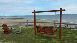 A wooden swing set and two Adirondack chairs overlooking a beach with a view of the ocean and distant landforms. - Ocean View Apartment Along Maine's Scenic Penobscot Bay (Belfast)