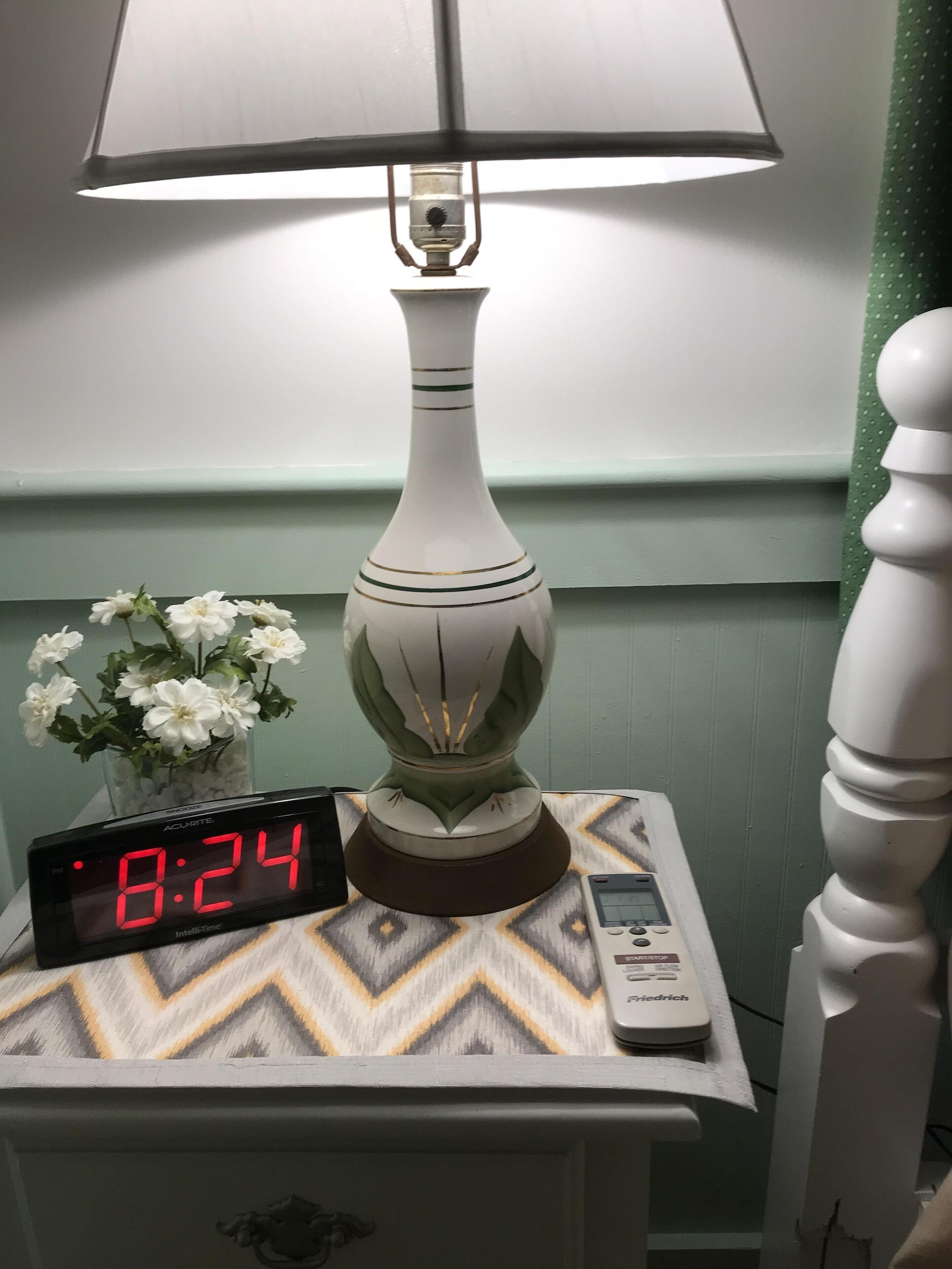 A bedside table with a lamp, a digital clock displaying 8:24, and a vase with white flowers.