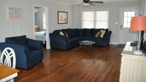A living room with a dark blue sectional sofa, a white armchair, and a coffee table. There is a ceiling fan, a framed picture on the wall, and a door with a window. - Ocean view, Pet Friendly house with private swimming pool! Sleeps 10. (North Myrtle Beach)