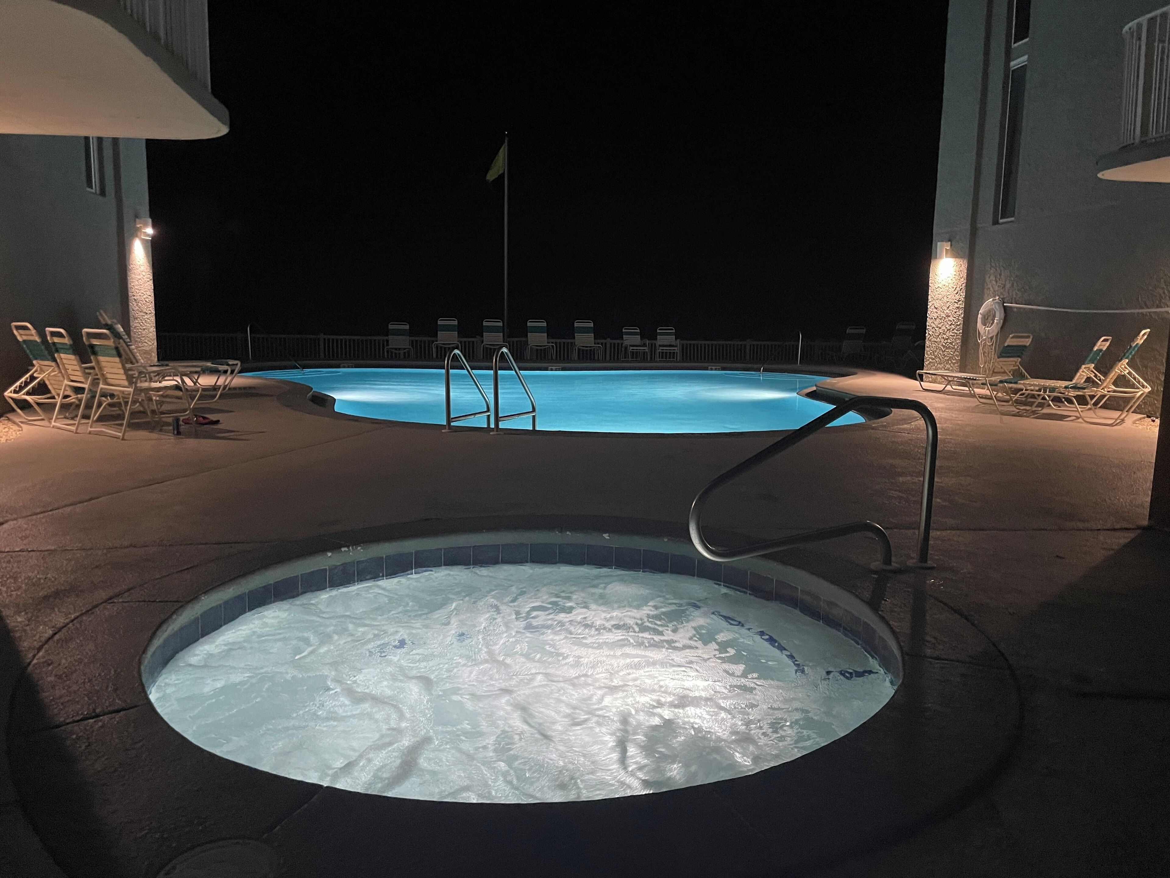 A swimming pool with lounge chairs and a hot tub at night.