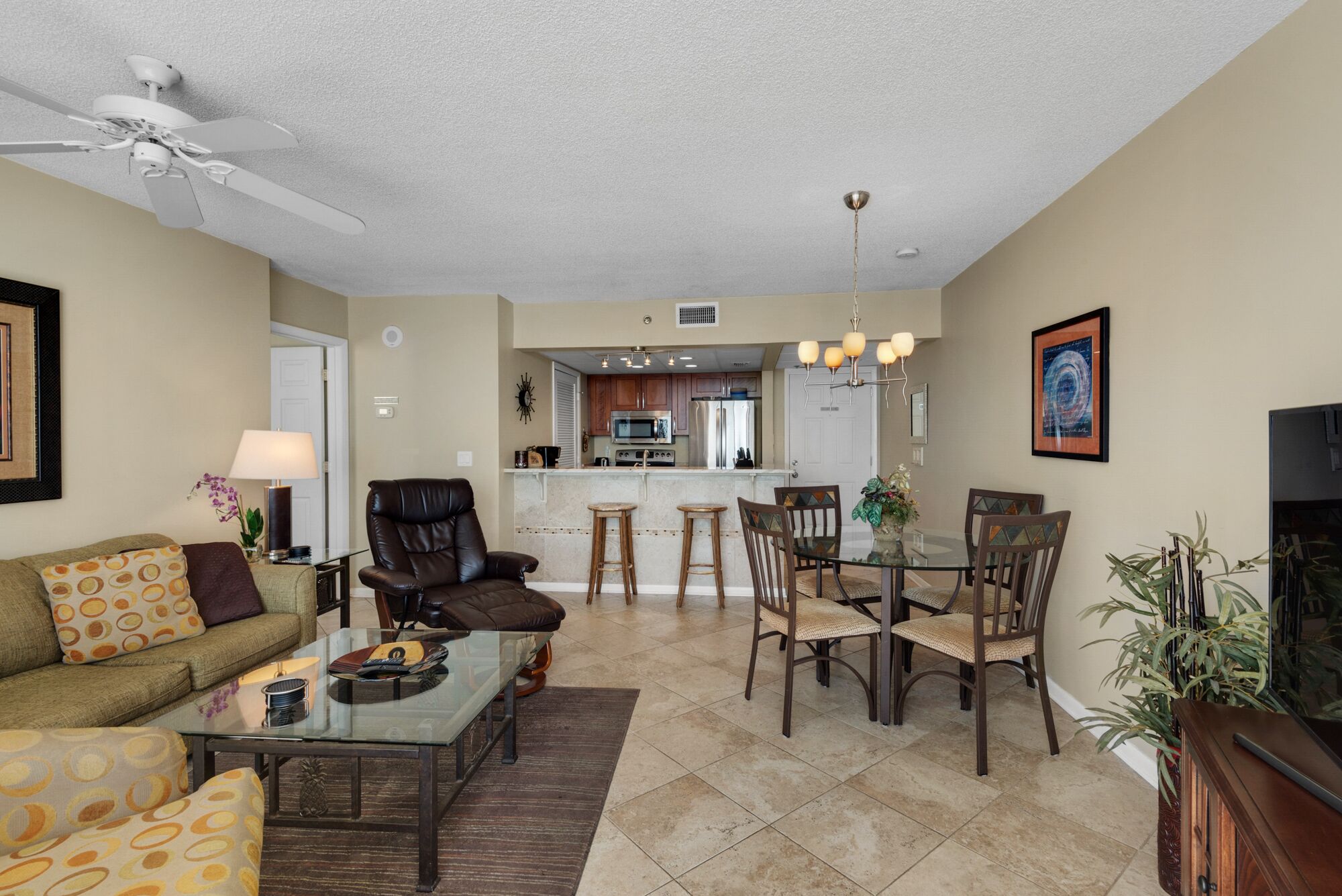 A living room with a sofa, coffee table, and chairs, a dining area with a table and chairs, a kitchen with appliances, and a ceiling fan.