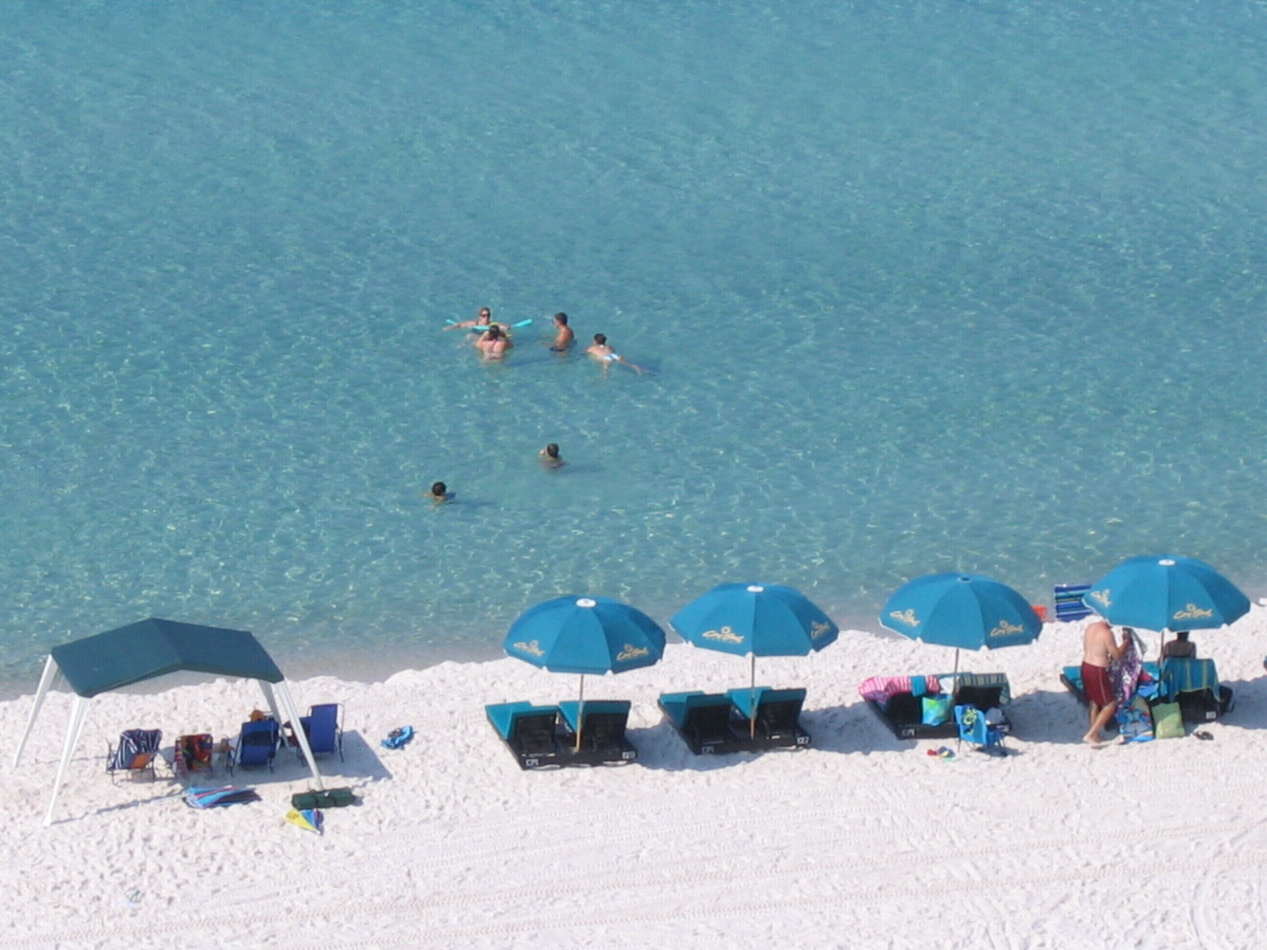Una playa con agua azul cristalina, gente nadando y varios sombrillas y tumbonas azules.