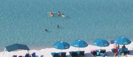 Una playa con agua azul cristalina, gente nadando y varios sombrillas y tumbonas azules.
