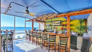 Un bar sulla spiaggia con vista sull'oceano, con sedie di legno, un bancone bar e una ventola a soffitto.
