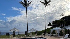 Ein Schwimmbad mit Liegestühlen und Sonnenschirmen, umgeben von Palmen und einem Zaun.