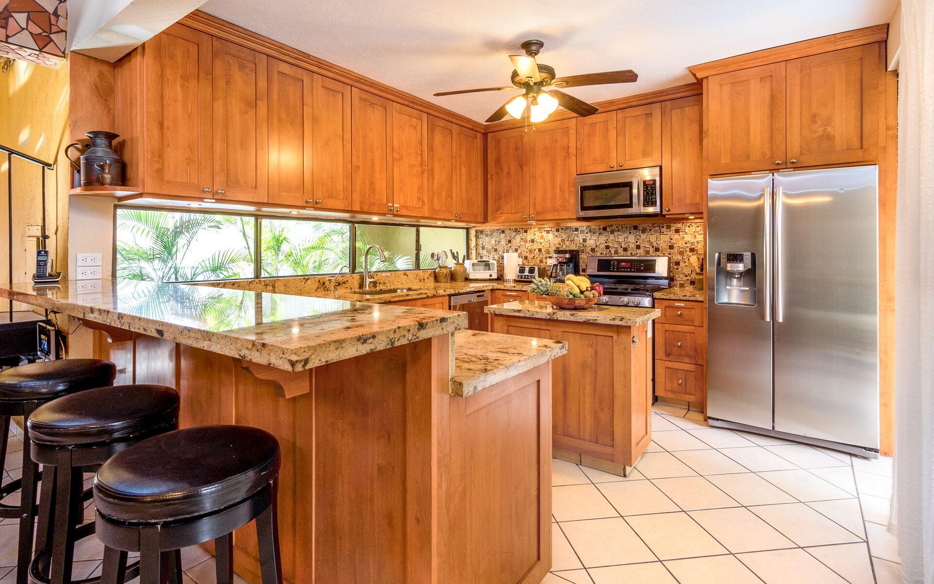 A kitchen with wooden cabinets, a central island with stools, a refrigerator, a microwave, and a ceiling fan.