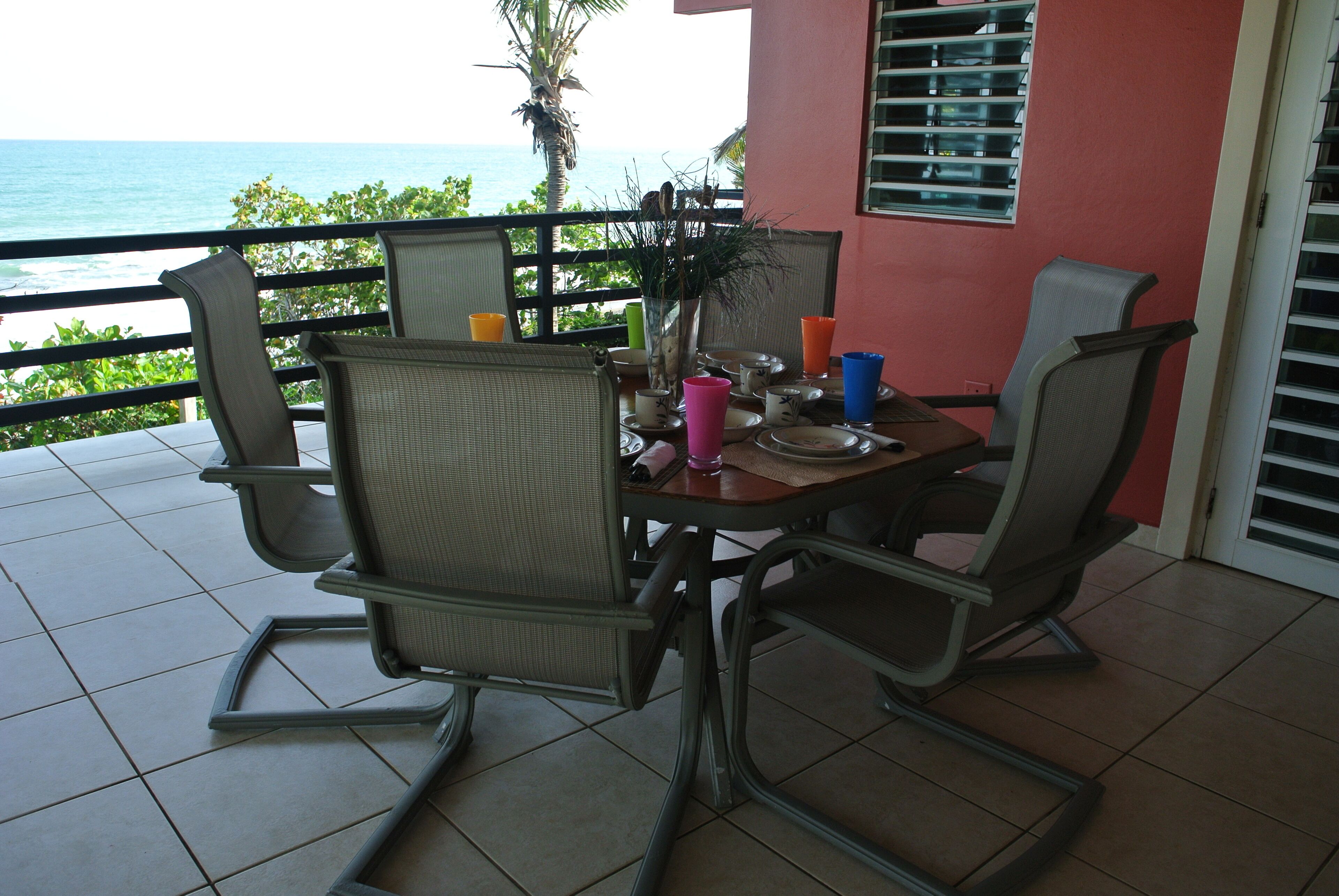 A dining area with a view of the ocean, equipped with wicker chairs and a table set with plates, cups, and a vase.