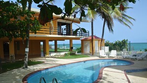 A pool area with a clear view of the ocean, a two-story building with balconies, and a small structure with a red roof.