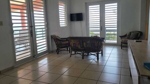 A room with a tiled floor, a dining table with chairs, a sofa, and a TV mounted on the wall.