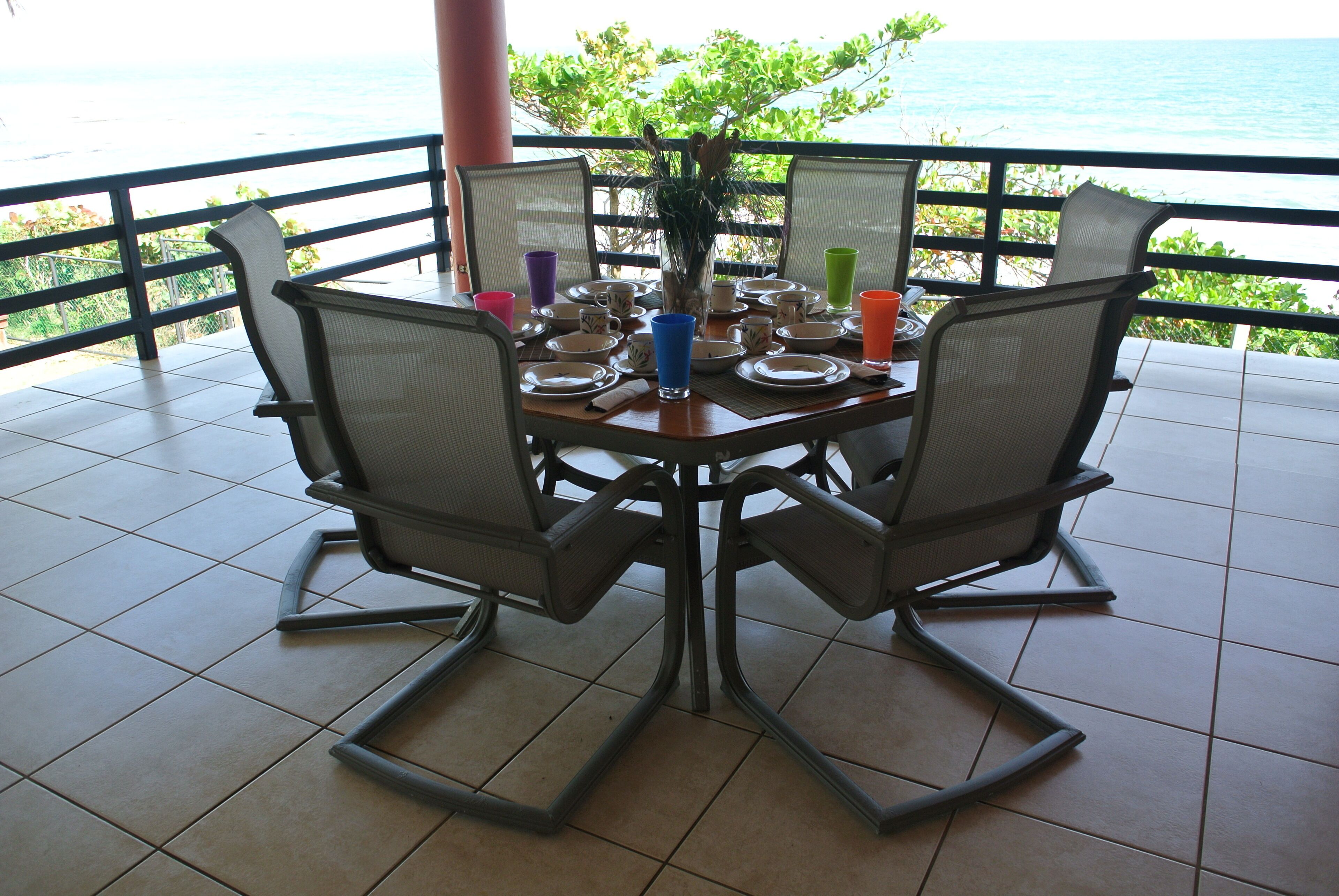 A dining table with chairs, a vase with flowers, and various cups and plates on a balcony with a view of the ocean.