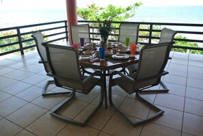 Een eettafel met stoelen, een vaas met bloemen, en verschillende kopjes en borden op een balkon met uitzicht op de oceaan.