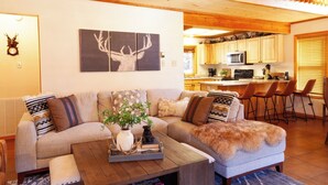 A cozy living room with a sofa, coffee table, and a decorative plant. A kitchen is visible in the background with wooden cabinets and a microwave. - Deer Mountain Lodge - High-End Home - In Town - Hot Tub - Free WiFi - Washer/Dry (Red River)
