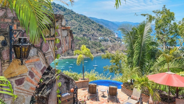 A patio with outdoor seating, a view of a bay with boats, and a mountainous backdrop.