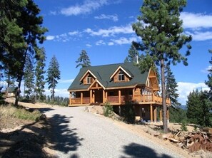 A log cabin with a prominent porch and a steep driveway, surrounded by tall pine trees. - Jack Pine Lodge:  Beautiful New Log Home (Cle Elum)