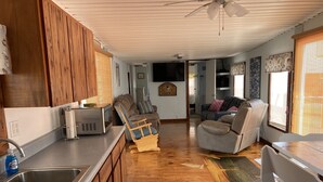 A spacious living area with a wooden floor, a kitchen with a sink and appliances, a ceiling fan, and a couch with a patterned cushion. - Natural High-Amazing Beach Getaway-Privacy, Easy Access, Kayaking, Boating (Cedar Island)