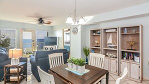 A living room with a dining table, chairs, a TV, and a bookshelf with nautical decorations. - Direct Gulf front View two bedroom, two bath totally refurbished. (Gulf Shores)
