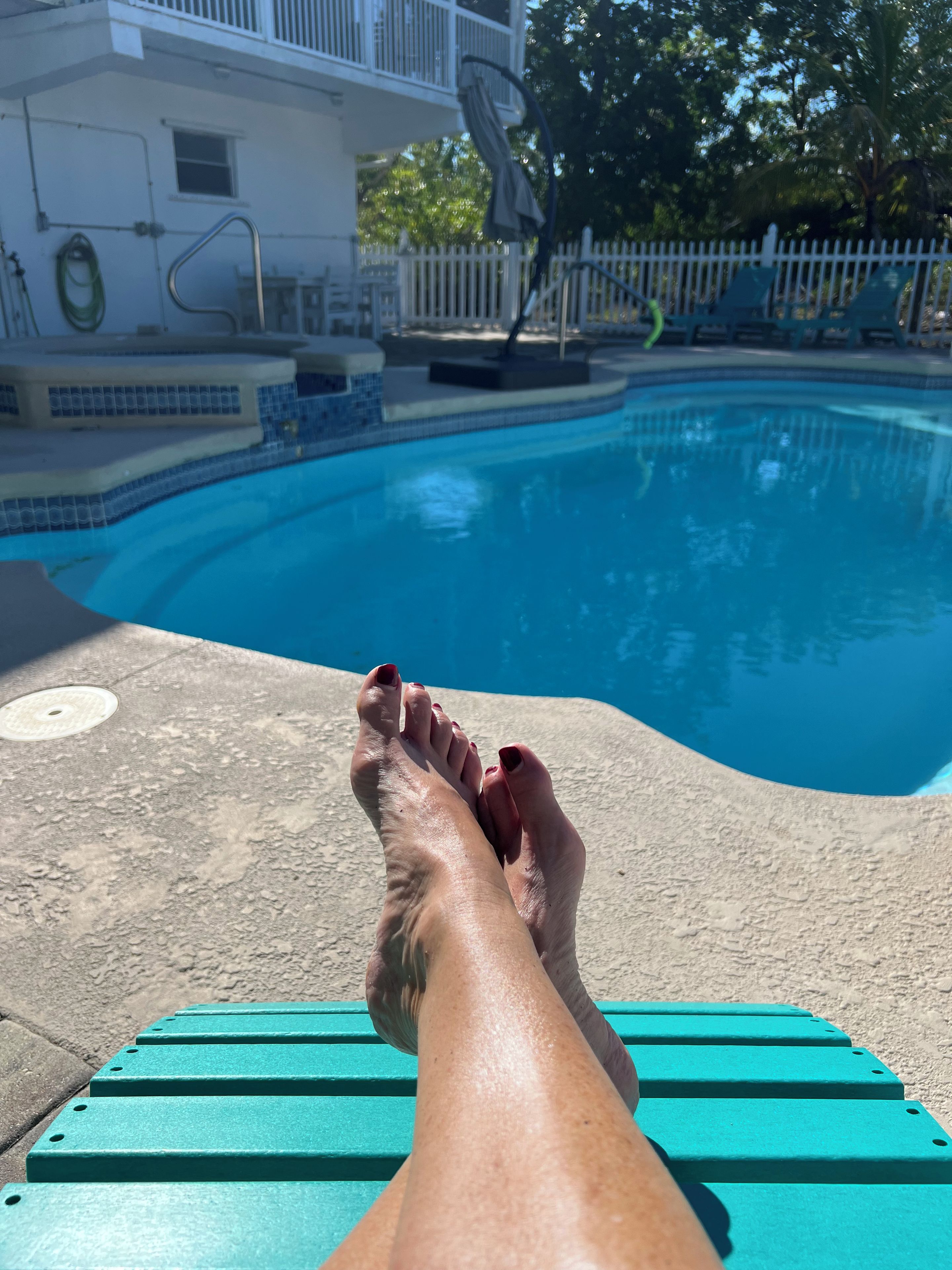 Vista de la piscina con agua azul cristalina, una cerca blanca y un edificio con balcón al fondo.