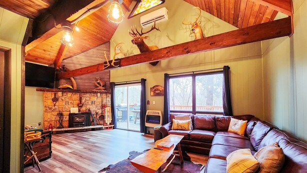 A rustic cabin interior with a wooden ceiling, stone fireplace, leather sofa, and a large window. - Secluded Pocono lodge, perfect for a weekend getaway with friends or family (East Stroudsburg)
