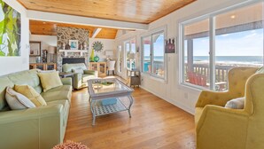 A spacious living room with a wooden ceiling, a stone fireplace, and large windows offering a view of the ocean. - "Ocean Kiss" - Oceanfront Home, 6 Bedrooms / 4 Bathrooms (Holden Beach)