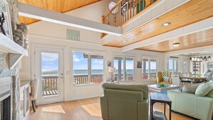 A spacious living room with a fireplace, a green sofa, and a view of the ocean through large windows. - "Ocean Kiss" - Oceanfront Home, 6 Bedrooms / 4 Bathrooms (Holden Beach)