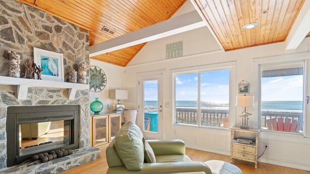 A living room with a stone fireplace, a green sofa, wooden flooring, and a view of the ocean through large windows. - "Ocean Kiss" - Oceanfront Home, 6 Bedrooms / 4 Bathrooms (Holden Beach)