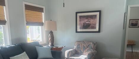 A living room with a sectional sofa, a patterned armchair, a white ottoman, a ceiling fan, and a framed picture on the wall.