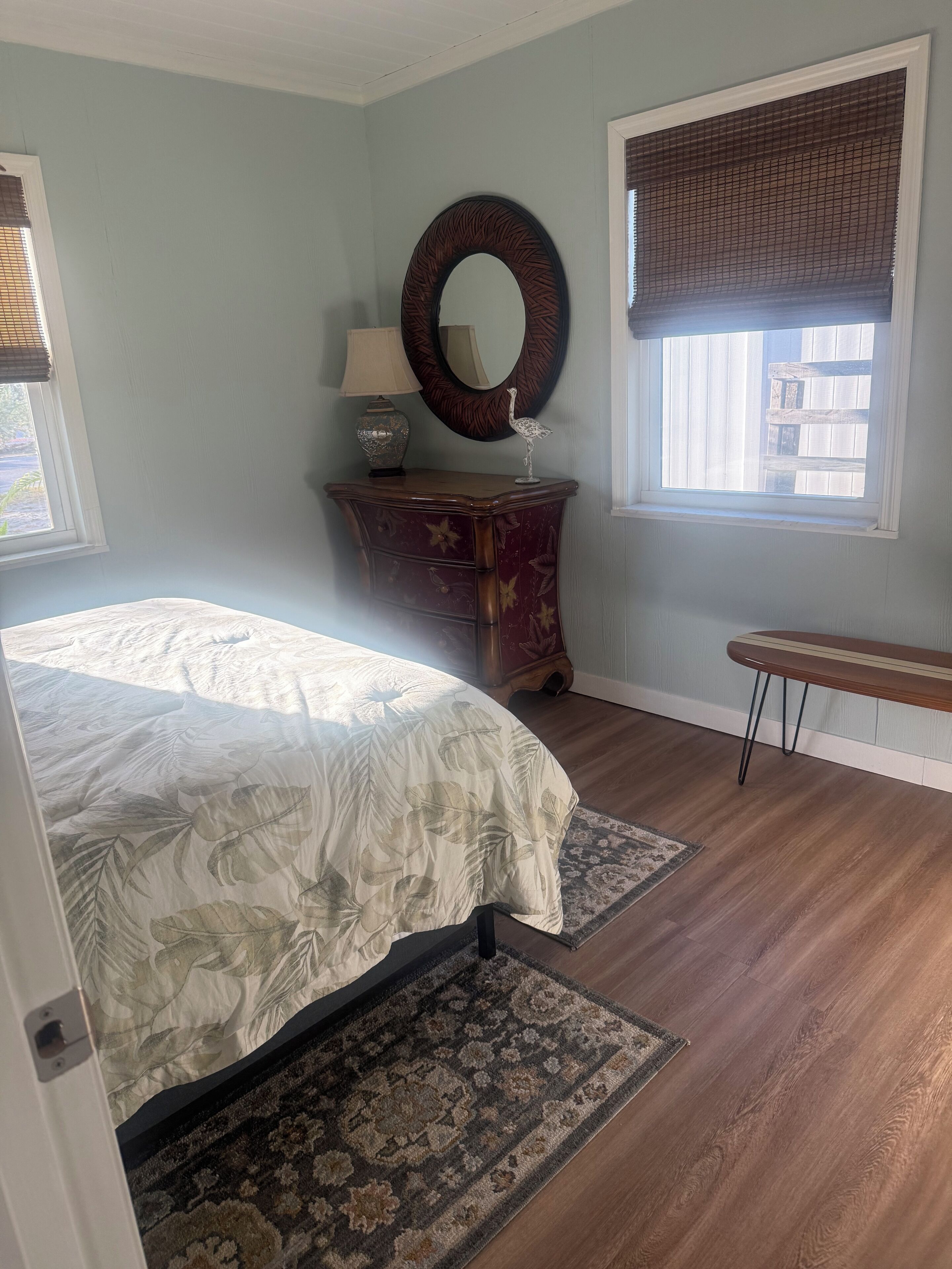 Une chambre à coucher comprenant un lit, une commode, un miroir, une fenêtre avec des stores et un banc.