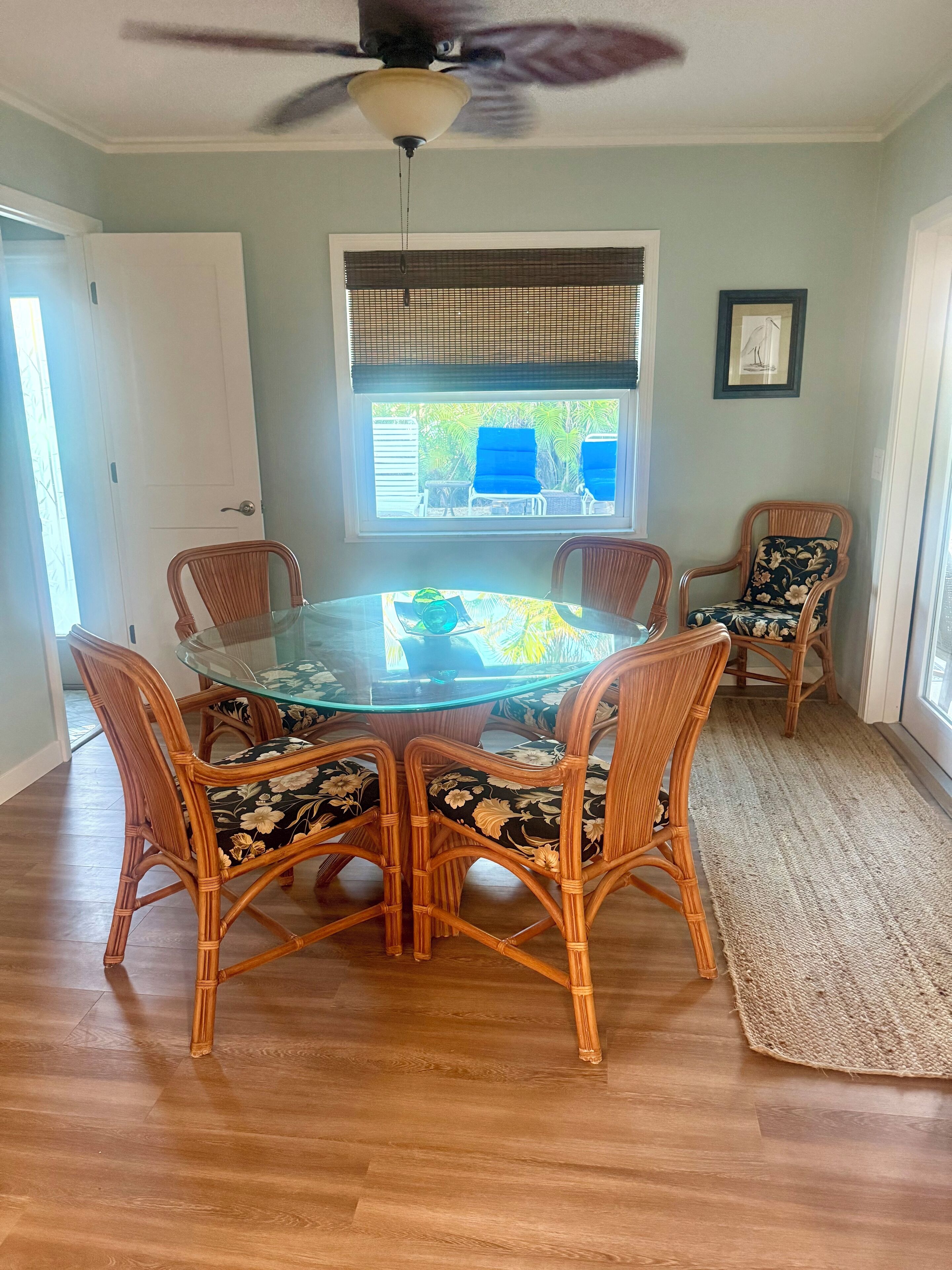 A dining area with a glass-top table surrounded by wicker chairs with floral upholstery, a ceiling fan, a window with blinds, and a framed picture on the wall.