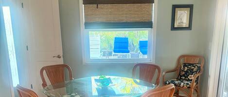 A dining area with a glass-top table surrounded by wicker chairs with floral upholstery, a ceiling fan, a window with blinds, and a framed picture on the wall.
