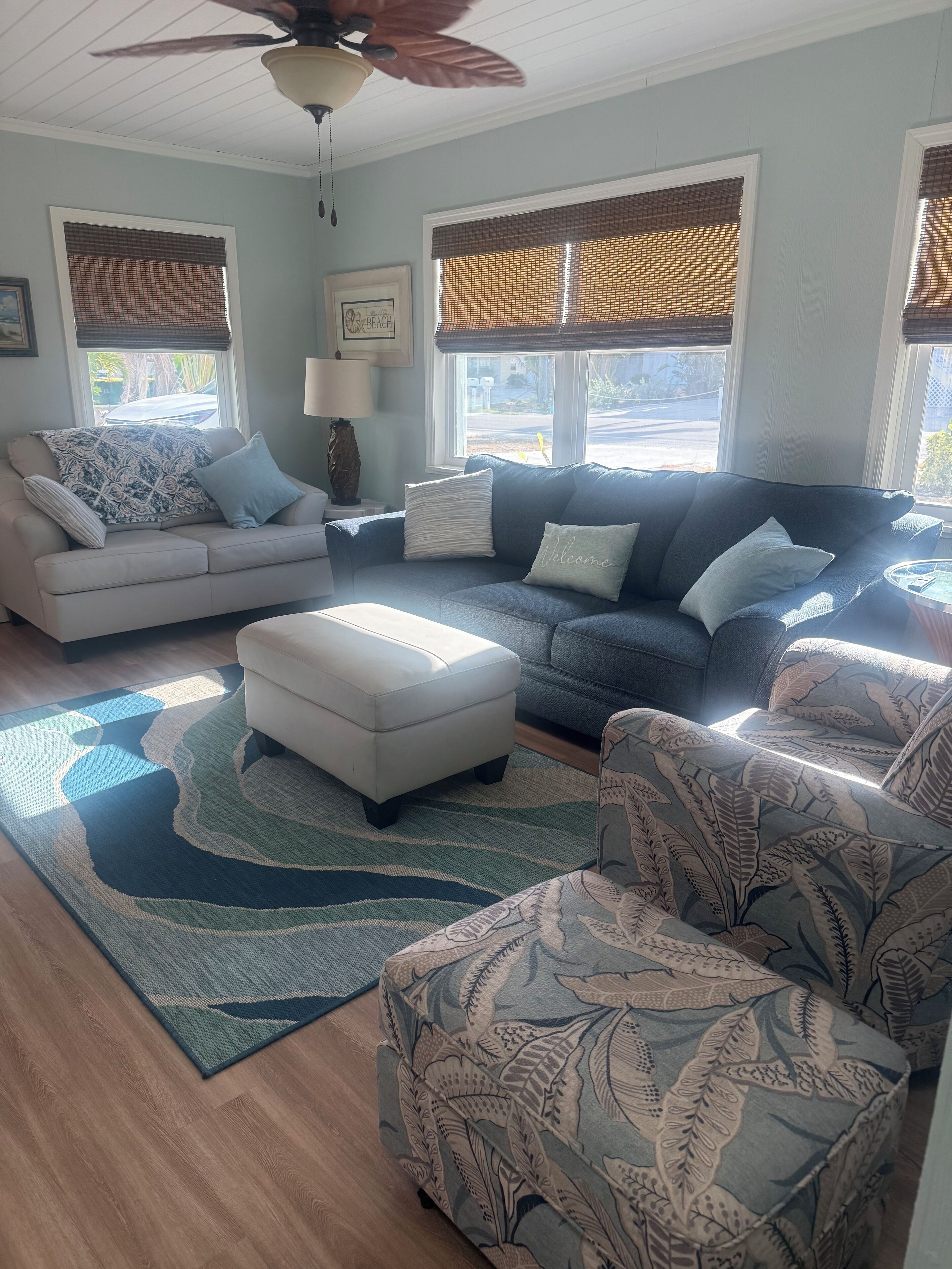 Una sala de estar con un sofá beige, un sofá azul, un sillón estampado y un reposapiés blanco. Hay dos ventanas con persianas, un ventilador de techo y una alfombra con motivos de hojas.